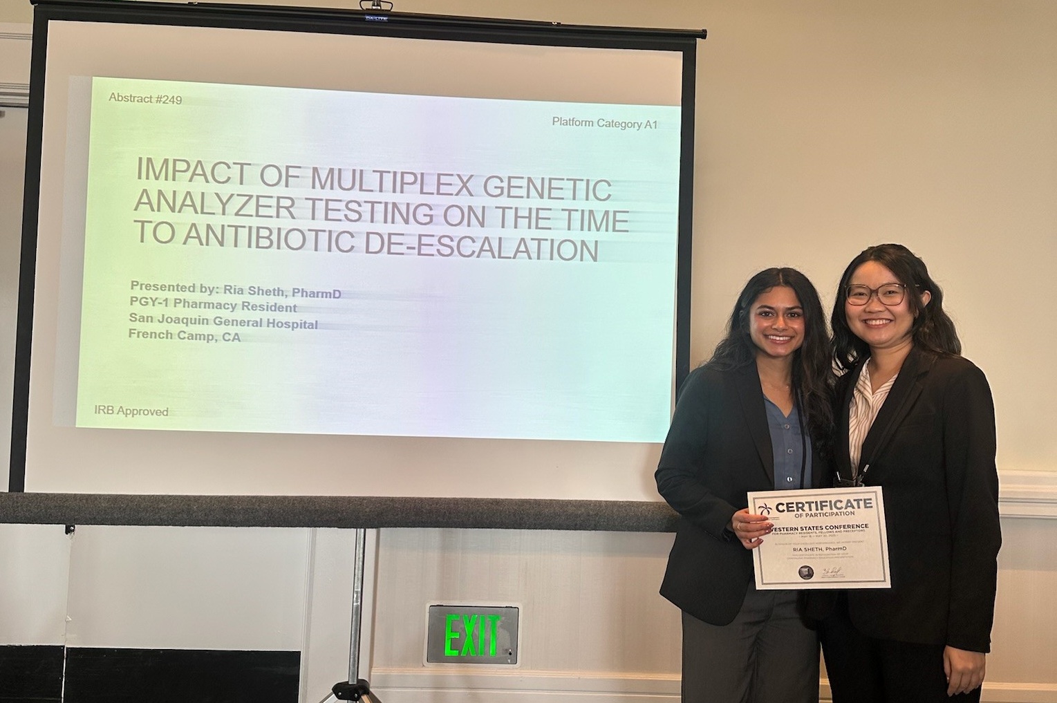 	
Two women stand smiling in front of a presentation slide titled Impact of Multiplex Genetic Analyzer Testing on the Time to Antibiotic De-escalation. One woman holds a certificate, and both appear to be at a professional event.